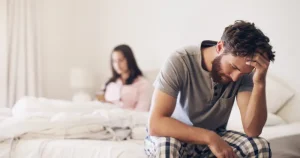 Distressed man sitting on the edge of a bed with his head in his hand while his partner sits apart in the background, suggesting relationship conflict and emotional distance.