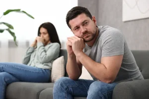 Worried couple sitting apart on a couch, with a thoughtful man leaning forward while his distressed partner sits in the background, showing emotional distance and relationship tension.