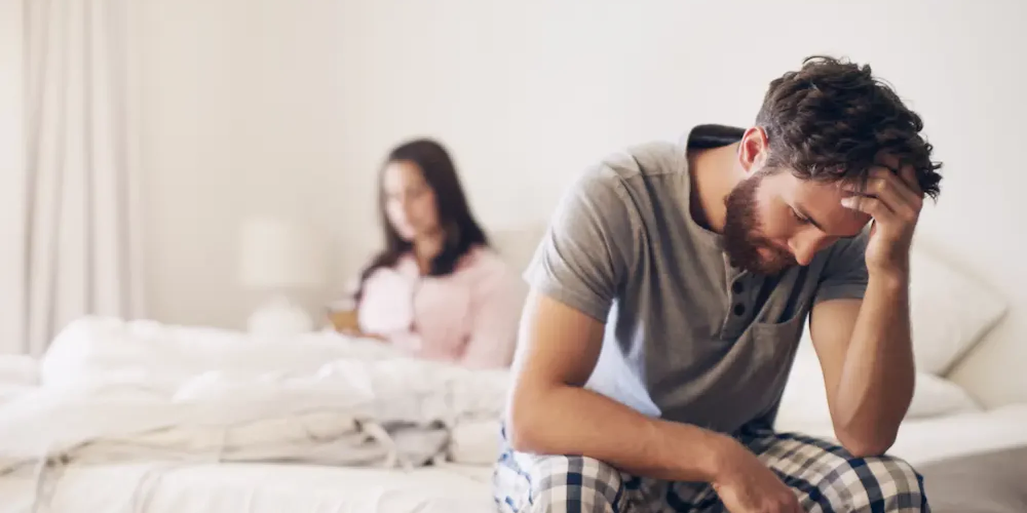 Distressed man sitting on the edge of a bed with his head in his hand while his partner sits apart in the background, suggesting relationship conflict and emotional distance.