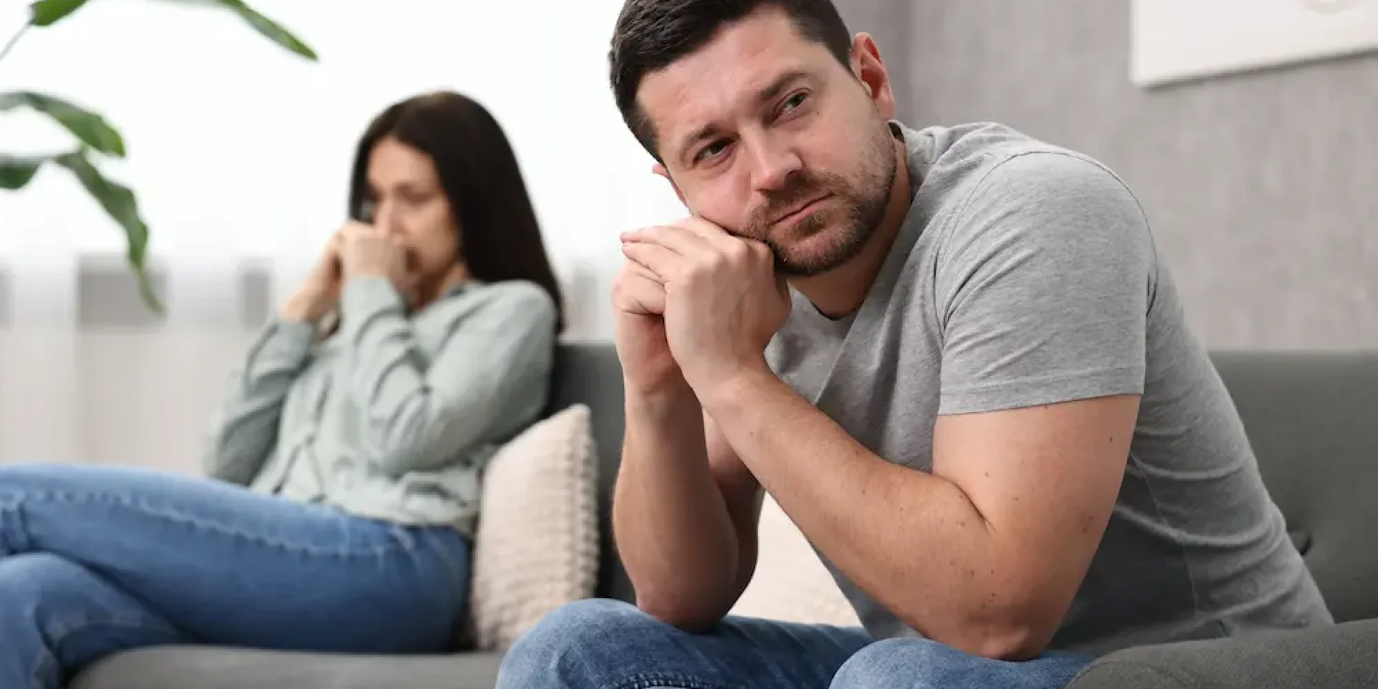 Worried couple sitting apart on a couch, with a thoughtful man leaning forward while his distressed partner sits in the background, showing emotional distance and relationship tension.